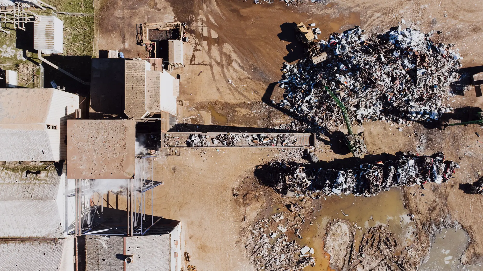 A recycling yard seen from directly above: sheds along one side, two long heaps of scrap metal and a conveyor across bare ground